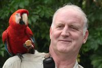 Bill Durham with a red parrot on his shoulder in front of a greenery background. in front of Photo by Claire Menke