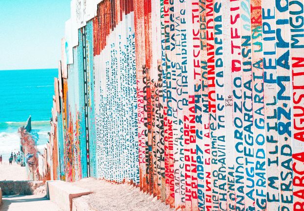 Image of bright lettering on the U.S.-Mexico border wall extending into a vibrant Pacific Ocean in the distance. Photo by Barbara Zandoval on Unsplash.